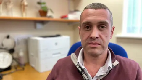 A doctor wearing a dark red top sitting at his desk