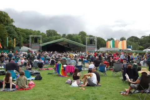 A crowd of people, many sitting on camping chairs, in front of an outdoor stage in a park. Festival flags in bright colours flutter near the stage.