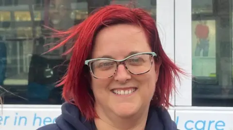 Andrew Turner/BBC Louise Frosdick standing next to the Apollo roadshow bus. She has shoulder -length red hair and is wearing glasses.  She is smiling. 