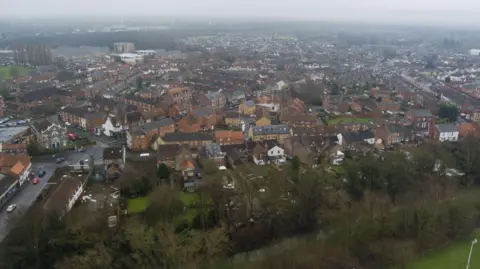 This is an overhead shot of Selby town centre, taken on a damp, cloudy day. Selby beck, which runs along the edge of the town centre, is in the foreground of the image. The historic town hall, with its distinctive white-painted wood and stonework, is to the left. The village of Thorpe Willoughby can be seen in the distance.