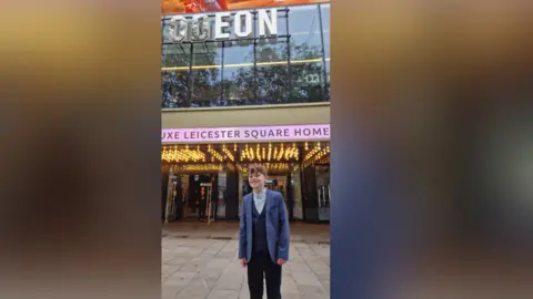 Nathan Lintern James stands in front of the Odeon in Leicester Square, London. He wears a blue jacket, navy waistcoat and trousers. 