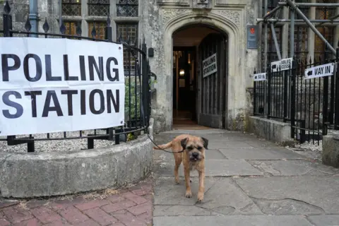 Carlos Jasso / AFP via Getty Images A small brown dog walks towards the camera next to a sign reading 'polling station' in front of a church in Lewes.