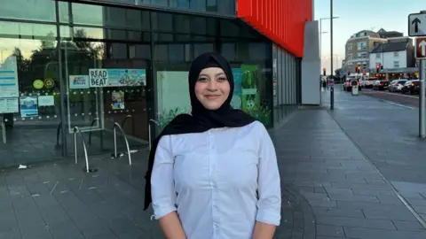 Leena is wearing a white shirt and is behind Slough high street.