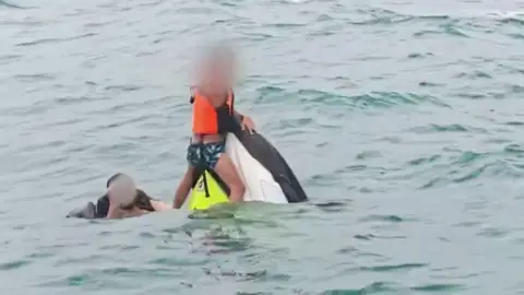 A boy wearing a lifejacket holds on to the top of a capsized jet ski as a man swims next to him in the sea