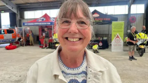 Kim Carey smiling at the camera. She is wearing glasses, a white jacket. In the background are two pitches including a blood bike and rescue group. 