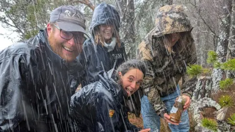 RZSS Four people are looking into the camera, smiling, one of them is holding a jar with hoverflies. It is pouring rain outside, they are standing in the forest.