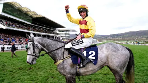 David Davies for The Jockey Club/PA Media Jockey smiling on top of a grey horse at Cheltenham racecourse with packed grandstands in the background. The jockey is wearing a yellow top with wide red bands on it