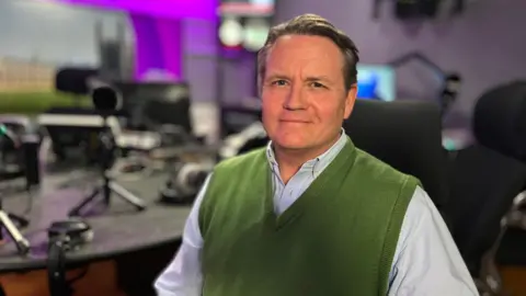 Martin Giles/BBC Duncan Bullivant wears a light blue shirt and green waistcoat. He stares at the camera and sits in front of a radio desk