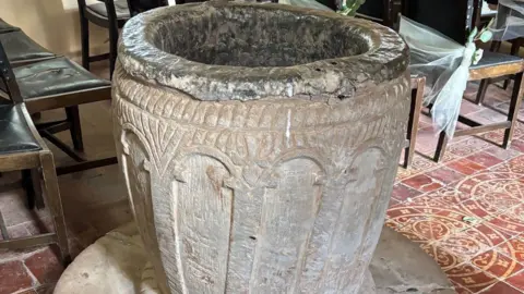 PA Media A round stone font in the shape of a barrel on a tiled floor with chairs behind it.