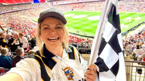 A woman holding a black and white racing flag and a football shirt stands in the stands of a large stadium. A football pitch can be seen behind her.
