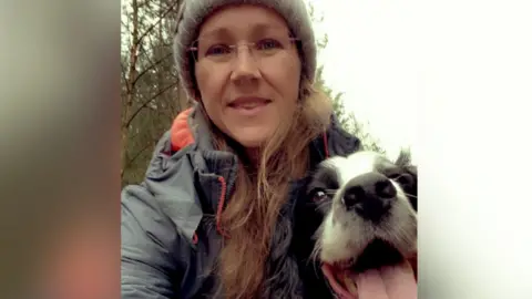 PA Media Mel Harrison wearing a hat and coat while standing with her dog in a wooded area. Her dog is a white and black spaniel and has its tongue hanging out.