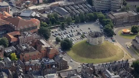 City of York Council The image shows a stone structure on top of a grassy mound in the center of a city area. Surrounding the mound are roads, parking spaces, and a mix of older brick buildings. A paved road encircles the mound, with vehicles visible along its perimeter.