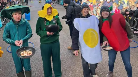Freya Cox/BBC A mixed group of people dressed in fancy dress pose holding frying pans with pancakes in them, on a busy street. From left to right, one man is dressed as green lizard, a woman is a sunflower, a younger woman is a fried egg and a fourth young woman is a tomato.
