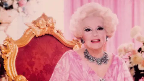 Cartland Promotions Barabara in her older years wearing pale makeup and with white-golden coiffed short hair in a perm. She wears a chunky silver sparkly necklace and matching earrings and pink chiffon blouse, sitting on a red and gilt throne with pink curtains behind her