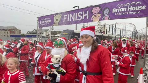 Blackpool Santa Dash A woman in Christmas tree-rimmed glasses holds a white dog in a red Santa suit as she joins hundreds of people dressed up in red Santa suits and hats, walking under the finish line banner, which has the Brian House Children's Hospice logo on it. Part of the Big One rollercoaster can be seen in the background.