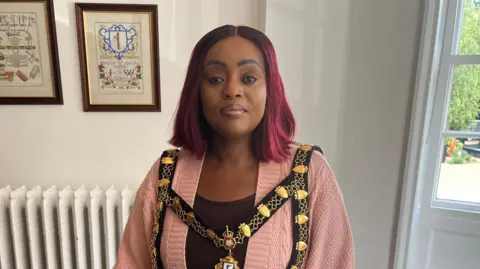 Tony Fisher/BBC Sally Kimondo, a black woman, wearing a pink cardigan and the mayoral chain over her shoulders looking into the camera. In the background is a wall with two pictures and a radiator