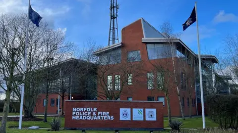 BBC Three-storey brick building with glass around the top floor. There are several antennae on a pole on top of the building, and another glass and brick building to the left. A sign showing "Norfolk Police and Fire Headquarters" is visible on grass in the foreground.