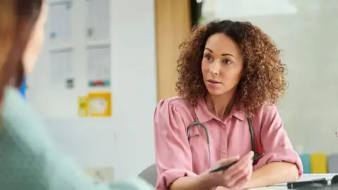 Getty Images A posed picture of a GP appointment - a woman wearing a pink shirt and with a stethoscope round her neck is sat at a desk talking to another woman who is out of focus on the left of the shot. The woman in the pink shirt has brown curly hair and is holding a pen in her hand. 