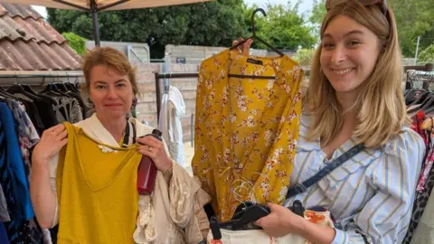Swap Society Two women hold up clothes and hangers in an outdoor setting, Both women are holding up a yellow tops and smiling. 