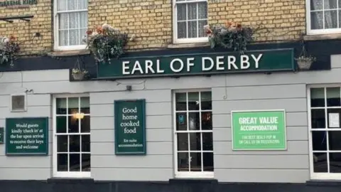 Robbie Kalus/BBC The Earl of Derby pub: a brown and grey building with white-framed windows. The words "Earl of Derby" are written in white capital letters on a green sign fixed to the front of the pub. Below the sign are signs advertising "good home cooked food" and "great value accommodation".