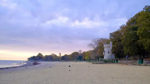 Martin Perry Ryde beach on the Isle of Wight. The wide sandy beach lined with trees with autumnal colours. There is a small castle-style stone tower on the shoreline.
