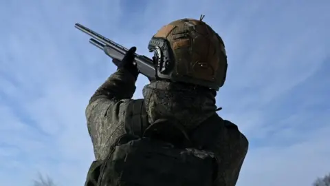 the back of a soldier in camouflage and helmet pointing a rifle towards a bright blue sky.