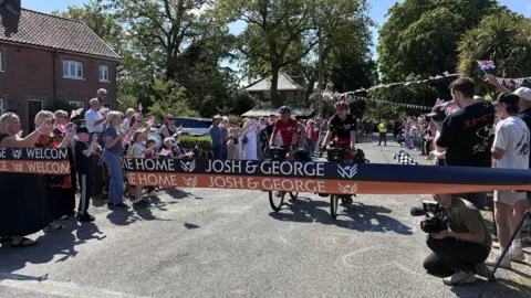 Jenny Kirk/BBC Josh and George Kohler are riding bicycles on a road. They are heading towards a piece of tape which says Josh & George on it. There are lots of people gathered either side of the road waving flags.
