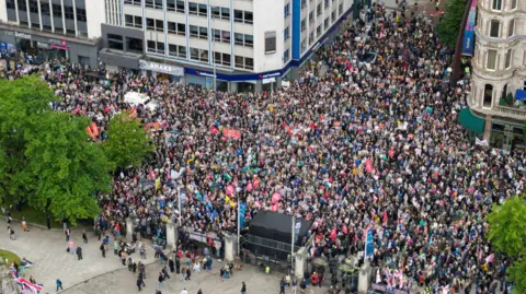 Reuters A drone image which shows thousands of people lining the streets around Belfast City Hall. Some are holding large flags, others are holding handmade posters. 