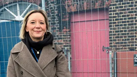 Clare Worden/BBC Jemma Curtis wears a cosy brown coat and black scarf. She has a blonde bob. She is standing in front of the brick annexe building, which has a red door.