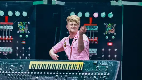 Sam Battle performing in a pink boiler suit. he is gesturing and stood next to a large keyboard with many buttons and dials on it. Large electronic equipment is visible in the background as stage decoration.