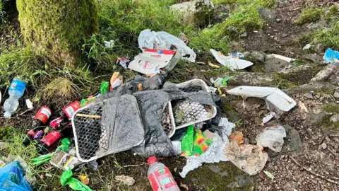 Central Beacons Mountain Rescue Team Three disposable barbecues are on the ground close to a tree, surrounded by other rubbish including plastic and glass bottles, food packaging and cigarette packets.