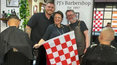 Two men and a woman, all wearing black, pose with a red and white flag - the colours of Sunderland AFC - in front of a wall which reads 'PJ's Barbershop'.