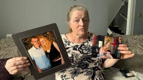 Margaret Bramall wearing a patterned dress holds up family photographs. The one in her right hand shows her daughter Lynsey and her partner, while she holds a photograph in her left hand showing her daughter smiling with her young son.