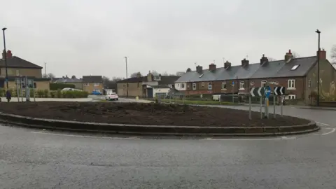 LDRS A roundabout that is covered in bare soil. Houses line the road leading up to the roundabout.