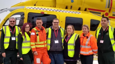 East Anglian Air Ambulance Steve Jones standing between East Anglian Air Ambulance staff, with two men and a woman on the left and two women and a man on the right. Behind him is a bright yellow ambulance. 