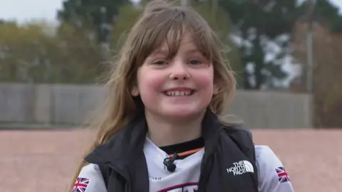 Close up image of Ella smiling at the camera. Ella has long brown hair with a fringe covering her forehead. She is wearing a white Exeter Eagles top with two Union Jacks on each arm. She has a black The North Face gilet on. She is sat on the BMX track. Green trees are in the background.