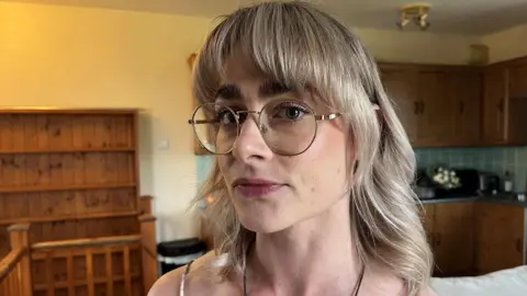 Georgina Cox with her head turned to the side, wearing metal-rimmed glasses. She has blonde hair and dark eyebrows with soft, natural makeup.