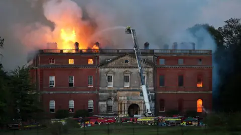 A three-storey stately Georgian home that is on fire. There are several fire engines at the base of the site and an aerial ladder platform is being used.