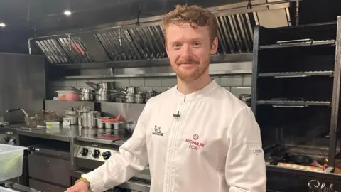 Ewan Waller is standing in the a restaurant kitchen, with pots and pans behind him. He smiles into the camera and wears a chefs apron that says 'Michelin 2026.' 