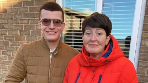 Joseph, a man in a brown coat and brown sunglasses. He has short cropped brown hair. He is standing next to Karen Denholm who is wearing a thick red jacket. She is smiling and has a short bobbed haircut. 