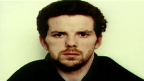 Gwent Police Mugshot of Jason Preece, who has curly brown hair and an auburn beard. He stares blankly into the camera and wears a dark jacket. 