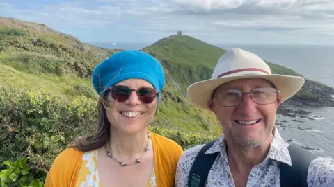 Lizzie Stroud A smiling selfie of Lizzy Stroud and husband Dave standing high on a cliff with the sea and hills behind them. She is wearing a yellow cardigan and blue beret and sunglasses. He is wearing a pale blue floral shirt a sunhat, glasses and backpack