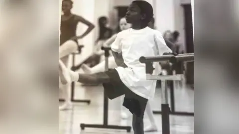 Nahum McLean/ Z Arts Stretford Young boy stands in a t-shirt and shorts against a ballet bar with one leg pointed straight forward. 