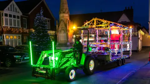 A small agricultural vehicle is lit  up in neon green lights and it is pulling a large trailer with a blow up Santa on it and Chrismas lights.