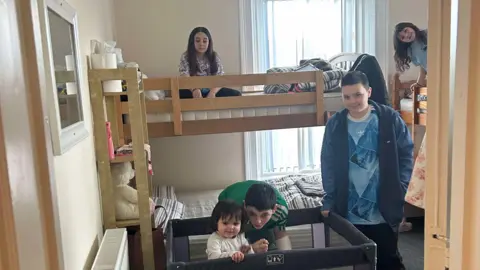 Louise McKillion Five children of varying ages in a small bedroom. Two girls are sitting in the top bunks of bunk beds. One boy is standing next to a travel cot, another boy is bent down next to a toddler standing in the cot.