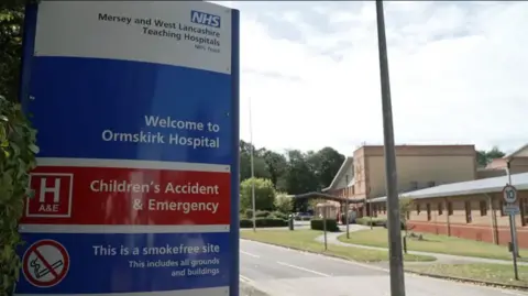 Close up of a sign which reads 'Welcome to Ormskirk Hospital' at the entry to the hospital building in the background on a clear day.