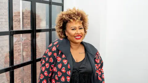 Emeli Sande smiles into the camera while standing indoors, next to a window. She is wearing a patterned jacket showing lots of hearts, and has brown hair.