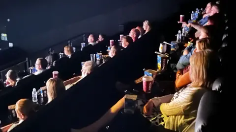 Emma Baugh/BBC Rows of people watching the screen inside a cinema theatre.