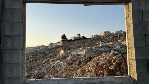 View through the empty window frame of a large pile of rubble in Nablus, with some buildings still standing in the distance