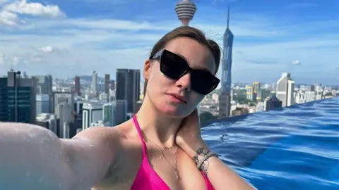 A selfie of Valeria Baigascina taken in a rooftop pool in Kuala Lumpur, with the striking skyline with tall towers behind her. Her long dark hair is tied back, she wears a pink bikini and dark sunglasses and a tattoo is visible on her wrist.